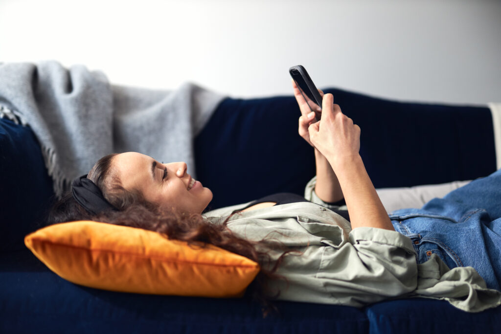 Profile view of young smiling mixed ethnicity woman lying on sofa at home checking social media on mobile phone